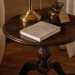 Un guéridon rond en bois sombre près d’un fauteuil, lampe et vase.